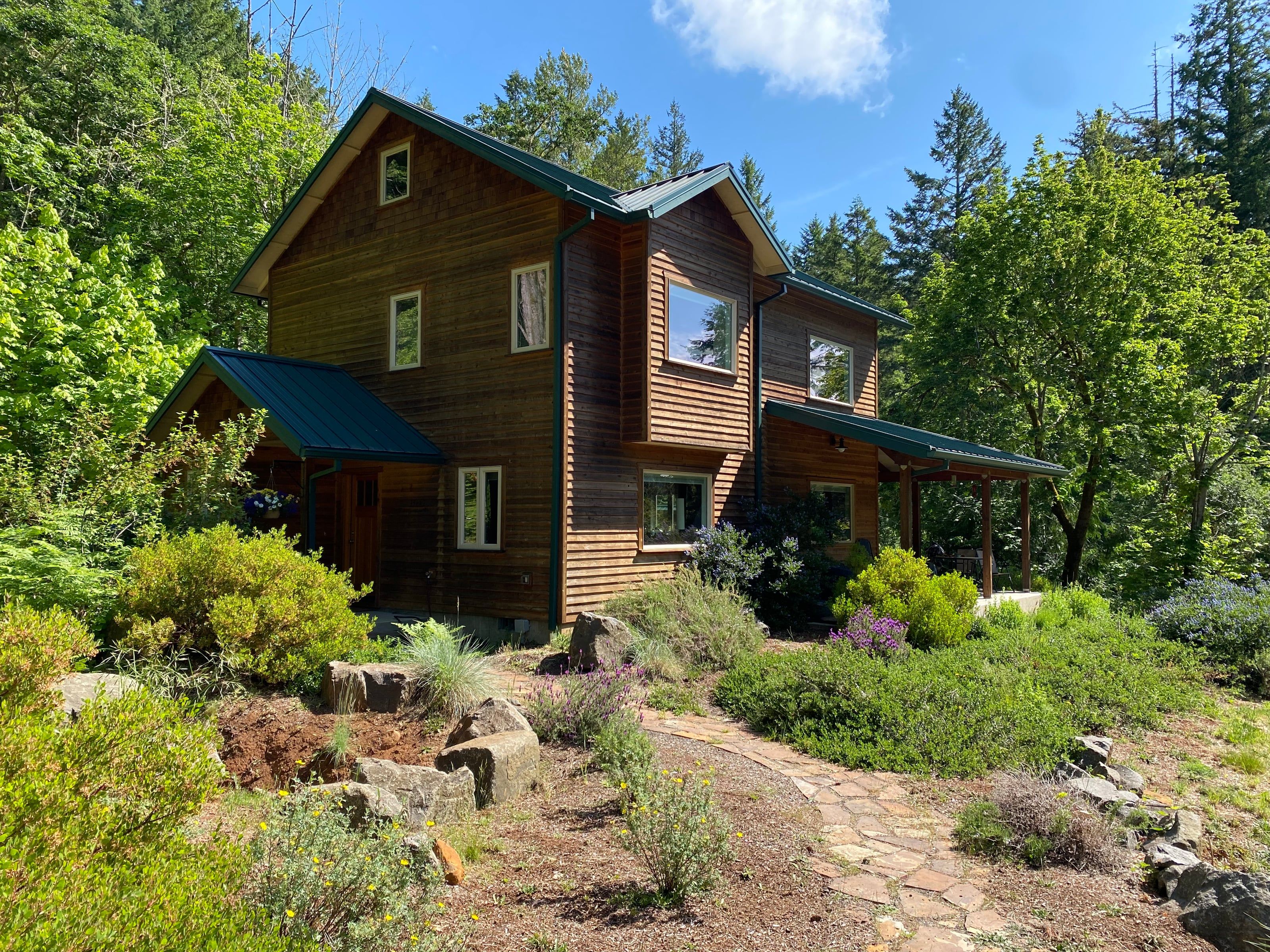 Cedar-clad two-story home with forest-green metal roofs behind rock-and-lavender landscaping, Douglas-firs beyond